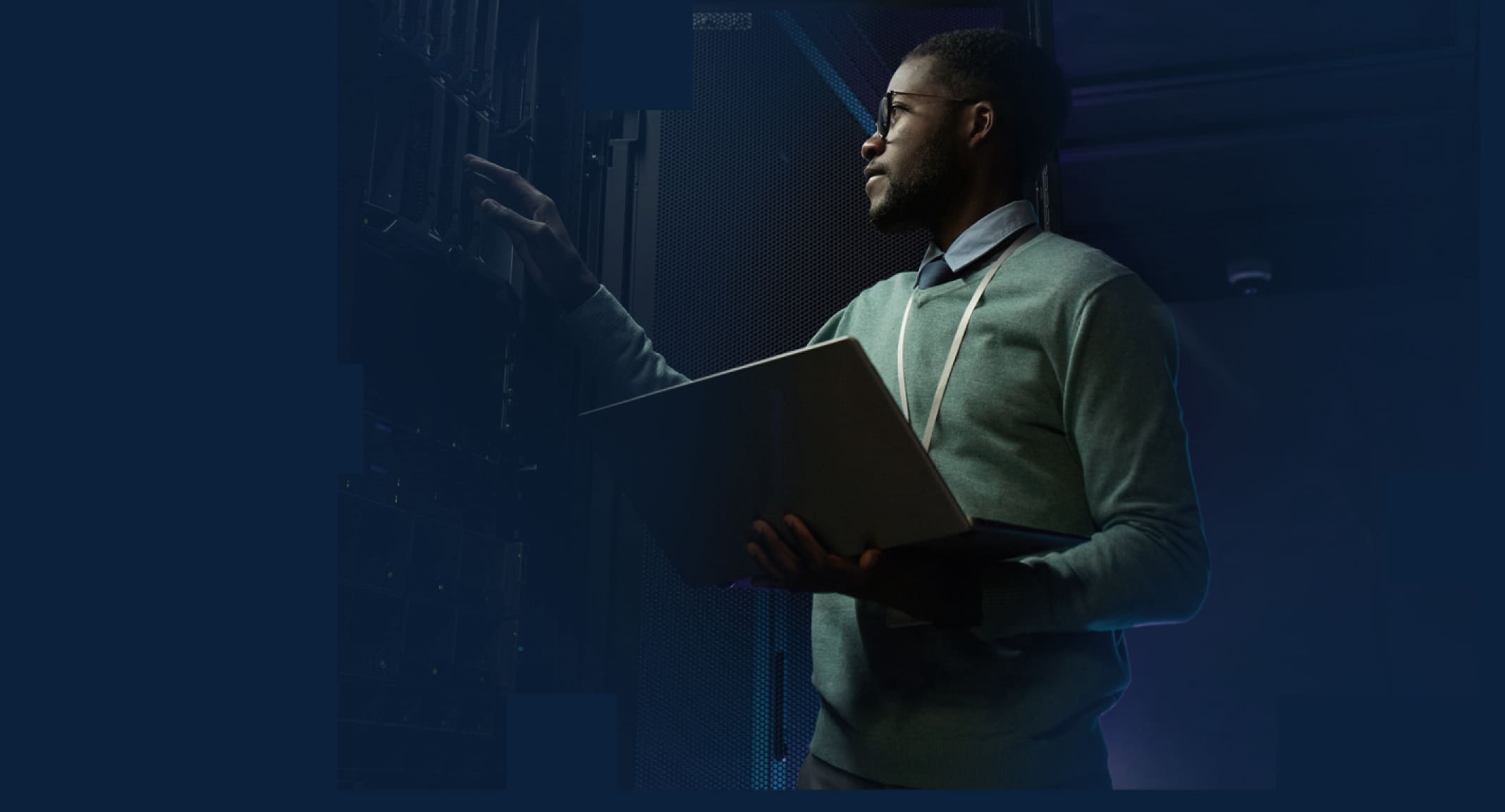 An IT professional wearing glasses working on a server rack with a badge around his neck and a laptop in hand